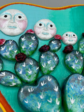 Load image into Gallery viewer, This handcrafted polymer clay wall art features a heart-shaped wood frame complimented with three smiling moon motifs.  Each moon symbolizing each of the realms of heavens, earth, and underworld. The complimentary color palette consists of cool sky hues, from a bright turquoise to a dark royal blue. The piece is further enhanced with hand sculpted cacti plants with blooming light and dark pink flowers.  The vibrant red and orange frame complements the cool-toned artwork.   H 7" x W 6 3/4” x D 1 1/4"