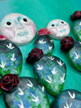 Load image into Gallery viewer, This handcrafted polymer clay wall art features a heart-shaped wood frame complimented with three smiling moon motifs.  Each moon symbolizing each of the realms of heavens, earth, and underworld. The complimentary color palette consists of cool sky hues, from a bright turquoise to a dark royal blue. The piece is further enhanced with hand sculpted cacti plants with blooming light and dark pink flowers.  The vibrant red and orange frame complements the cool-toned artwork.   H 7" x W 6 3/4” x D 1 1/4"