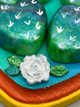 Load image into Gallery viewer, This handcrafted polymer clay wall art features a heart-shaped wood frame complimented with three smiling moon motifs.  Each moon symbolizing each of the realms of heavens, earth, and underworld. The complimentary color palette consists of cool sky hues, from a bright turquoise to a dark royal blue. The piece is further enhanced with hand sculpted cacti plants with blooming light and dark pink flowers.  The vibrant red and orange frame complements the cool-toned artwork.   H 7" x W 6 3/4” x D 1 1/4"