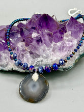 Load image into Gallery viewer, This Dark Gray-blue Agate Geode Sliced Stone Chip Necklace is a unique, eye-catching accessory to elevate your jewelry collection. The bold pendant features a striking gray-blue agate geode surrounded by eye-catching, shimmery dark-purple glass beads with beautiful iridescence that reflects hues of blue, pink, gold, and green. It's a perfect boho chic accent to update any look.   Measurements:   Necklace is 18” in length - pendant is 1 1/4” Height