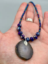 Load image into Gallery viewer, This Dark Gray-blue Agate Geode Sliced Stone Chip Necklace is a unique, eye-catching accessory to elevate your jewelry collection. The bold pendant features a striking gray-blue agate geode surrounded by eye-catching, shimmery dark-purple glass beads with beautiful iridescence that reflects hues of blue, pink, gold, and green. It's a perfect boho chic accent to update any look.   Measurements:   Necklace is 18” in length - pendant is 1 1/4” Height