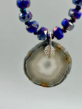 Load image into Gallery viewer, This Dark Gray-blue Agate Geode Sliced Stone Chip Necklace is a unique, eye-catching accessory to elevate your jewelry collection. The bold pendant features a striking gray-blue agate geode surrounded by eye-catching, shimmery dark-purple glass beads with beautiful iridescence that reflects hues of blue, pink, gold, and green. It's a perfect boho chic accent to update any look.   Measurements:   Necklace is 18” in length - pendant is 1 1/4” Height