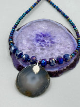 Load image into Gallery viewer, This Dark Gray-blue Agate Geode Sliced Stone Chip Necklace is a unique, eye-catching accessory to elevate your jewelry collection. The bold pendant features a striking gray-blue agate geode surrounded by eye-catching, shimmery dark-purple glass beads with beautiful iridescence that reflects hues of blue, pink, gold, and green. It's a perfect boho chic accent to update any look.   Measurements:   Necklace is 18” in length - pendant is 1 1/4” Height