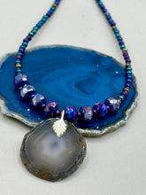 Load image into Gallery viewer, This Dark Gray-blue Agate Geode Sliced Stone Chip Necklace is a unique, eye-catching accessory to elevate your jewelry collection. The bold pendant features a striking gray-blue agate geode surrounded by eye-catching, shimmery dark-purple glass beads with beautiful iridescence that reflects hues of blue, pink, gold, and green. It's a perfect boho chic accent to update any look.   Measurements:   Necklace is 18” in length - pendant is 1 1/4” Height