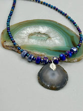 Load image into Gallery viewer, This Dark Gray-blue Agate Geode Sliced Stone Chip Necklace is a unique, eye-catching accessory to elevate your jewelry collection. The bold pendant features a striking gray-blue agate geode surrounded by eye-catching, shimmery dark-purple glass beads with beautiful iridescence that reflects hues of blue, pink, gold, and green. It's a perfect boho chic accent to update any look.   Measurements:   Necklace is 18” in length - pendant is 1 1/4” Height