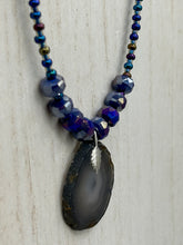 Load image into Gallery viewer, This Dark Gray-blue Agate Geode Sliced Stone Chip Necklace is a unique, eye-catching accessory to elevate your jewelry collection. The bold pendant features a striking gray-blue agate geode surrounded by eye-catching, shimmery dark-purple glass beads with beautiful iridescence that reflects hues of blue, pink, gold, and green. It's a perfect boho chic accent to update any look.   Measurements:   Necklace is 18” in length - pendant is 1 1/4” Height