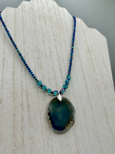 Load image into Gallery viewer, This Dark Blue Agate Geode Sliced Stone Chip Necklace is a luxurious addition to any jewelry collection. It features bold, eye-catching dark blue agate geode in an earthy outline, suspended on a unique glass beaded necklace that contains a mixture of blue and purple glass beads with a beautiful iridescence. This necklace is the perfect boho chic accent to take any look to the next level.   Measurements:    Necklace is 18” in length - pendant is 1 3/4” Height