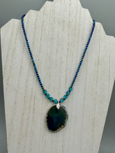 Load image into Gallery viewer, This Dark Blue Agate Geode Sliced Stone Chip Necklace is a luxurious addition to any jewelry collection. It features bold, eye-catching dark blue agate geode in an earthy outline, suspended on a unique glass beaded necklace that contains a mixture of blue and purple glass beads with a beautiful iridescence. This necklace is the perfect boho chic accent to take any look to the next level.   Measurements:    Necklace is 18” in length - pendant is 1 3/4” Height