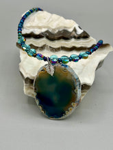Load image into Gallery viewer, This Dark Blue Agate Geode Sliced Stone Chip Necklace is a luxurious addition to any jewelry collection. It features bold, eye-catching dark blue agate geode in an earthy outline, suspended on a unique glass beaded necklace that contains a mixture of blue and purple glass beads with a beautiful iridescence. This necklace is the perfect boho chic accent to take any look to the next level.   Measurements:    Necklace is 18” in length - pendant is 1 3/4” Height