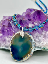 Load image into Gallery viewer, This Dark Blue Agate Geode Sliced Stone Chip Necklace is a luxurious addition to any jewelry collection. It features bold, eye-catching dark blue agate geode in an earthy outline, suspended on a unique glass beaded necklace that contains a mixture of blue and purple glass beads with a beautiful iridescence. This necklace is the perfect boho chic accent to take any look to the next level.   Measurements:    Necklace is 18” in length - pendant is 1 3/4” Height