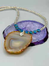 Load image into Gallery viewer, This handcrafted necklace is perfect for anyone who loves an earthy, bohemian look. Crafted with a unique strand of gold and clear beading, blue glass beads with iridescent hues, and natural banded agate geode, this piece adds style and luxury to any ensemble. Measurements: Necklace is 18” in length - pendant is 2” Height