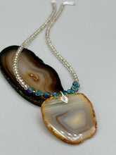 Load image into Gallery viewer, This handcrafted necklace is perfect for anyone who loves an earthy, bohemian look. Crafted with a unique strand of gold and clear beading, blue glass beads with iridescent hues, and natural banded agate geode, this piece adds style and luxury to any ensemble. Measurements: Necklace is 18” in length - pendant is 2” Height