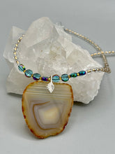 Load image into Gallery viewer, This handcrafted necklace is perfect for anyone who loves an earthy, bohemian look. Crafted with a unique strand of gold and clear beading, blue glass beads with iridescent hues, and natural banded agate geode, this piece adds style and luxury to any ensemble. Measurements: Necklace is 18” in length - pendant is 2” Height