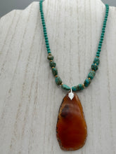 Load image into Gallery viewer, This unique and luxurious Amber Tone Agate Geode Sliced Stone Chip Necklace is a one-of-a-kind statement piece crafted from natural gemstones. The necklace features an amber-tone banded geode, suspended from a strand of blue and green turquoise and gold glass beads, and blue-green magnesite stone beads. This stylish boho-chic accessory is perfect for any casual or formal occasion. Measurements: Necklace is 18” in length - pendant is 2 1/2” Height