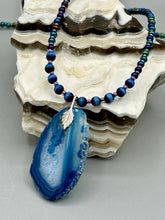 Load image into Gallery viewer, This Vibrant Royal Blue Agate Geode Sliced Stone Chip Necklace is a bold and beautiful statement piece. It features a natural gemstone pendant in a blend of light and dark blue tones, surrounded by glass beads in rich hues of blue, purple, gold, and green. This chic, boho-inspired necklace is completed by 10 blue cats eye beads and a silver leaf bail. Measurements: Necklace is 18” in length - pendant is 2 3/4” Height