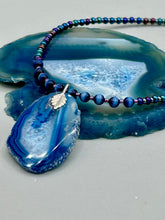 Load image into Gallery viewer, This Vibrant Royal Blue Agate Geode Sliced Stone Chip Necklace is a bold and beautiful statement piece. It features a natural gemstone pendant in a blend of light and dark blue tones, surrounded by glass beads in rich hues of blue, purple, gold, and green. This chic, boho-inspired necklace is completed by 10 blue cats eye beads and a silver leaf bail. Measurements: Necklace is 18” in length - pendant is 2 3/4” Height