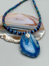 Load image into Gallery viewer, This Vibrant Royal Blue Agate Geode Sliced Stone Chip Necklace is a bold and beautiful statement piece. It features a natural gemstone pendant in a blend of light and dark blue tones, surrounded by glass beads in rich hues of blue, purple, gold, and green. This chic, boho-inspired necklace is completed by 10 blue cats eye beads and a silver leaf bail. Measurements: Necklace is 18” in length - pendant is 2 3/4” Height