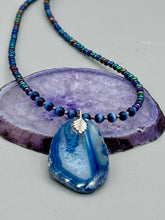 Load image into Gallery viewer, This Vibrant Royal Blue Agate Geode Sliced Stone Chip Necklace is a bold and beautiful statement piece. It features a natural gemstone pendant in a blend of light and dark blue tones, surrounded by glass beads in rich hues of blue, purple, gold, and green. This chic, boho-inspired necklace is completed by 10 blue cats eye beads and a silver leaf bail. Measurements: Necklace is 18” in length - pendant is 2 3/4” Height