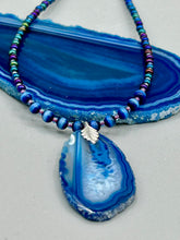 Load image into Gallery viewer, This Vibrant Royal Blue Agate Geode Sliced Stone Chip Necklace is a bold and beautiful statement piece. It features a natural gemstone pendant in a blend of light and dark blue tones, surrounded by glass beads in rich hues of blue, purple, gold, and green. This chic, boho-inspired necklace is completed by 10 blue cats eye beads and a silver leaf bail. Measurements: Necklace is 18” in length - pendant is 2 3/4” Height