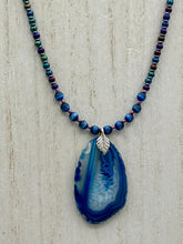 Load image into Gallery viewer, This Vibrant Royal Blue Agate Geode Sliced Stone Chip Necklace is a bold and beautiful statement piece. It features a natural gemstone pendant in a blend of light and dark blue tones, surrounded by glass beads in rich hues of blue, purple, gold, and green. This chic, boho-inspired necklace is completed by 10 blue cats eye beads and a silver leaf bail. Measurements: Necklace is 18” in length - pendant is 2 3/4” Height