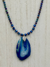 Load image into Gallery viewer, This Vibrant Royal Blue Agate Geode Sliced Stone Chip Necklace is a bold and beautiful statement piece. It features a natural gemstone pendant in a blend of light and dark blue tones, surrounded by glass beads in rich hues of blue, purple, gold, and green. This chic, boho-inspired necklace is completed by 10 blue cats eye beads and a silver leaf bail. Measurements: Necklace is 18” in length - pendant is 2 3/4” Height
