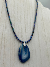Load image into Gallery viewer, This Vibrant Royal Blue Agate Geode Sliced Stone Chip Necklace is a bold and beautiful statement piece. It features a natural gemstone pendant in a blend of light and dark blue tones, surrounded by glass beads in rich hues of blue, purple, gold, and green. This chic, boho-inspired necklace is completed by 10 blue cats eye beads and a silver leaf bail. Measurements: Necklace is 18” in length - pendant is 2 3/4” Height
