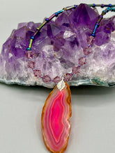 Load image into Gallery viewer, Make a bold style statement with this eye-catching Soft Pink and Amber Agate Geode Sliced Stone Chip Necklace. Featuring a vibrant soft pink and amber tone agate geode pendant surrounded by dark blue glass beads with iridescent tones of purple, blue, green, pink and gold. This bohemian-style gemstone necklace is the perfect way to add a unique touch of shabby boho chic to any look.   Measurements:    Necklace is 18” in length - pendant is 2 1/4” Height
