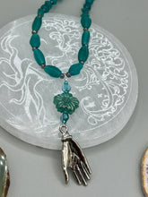 Load image into Gallery viewer, Bring the beauty of nature to your wardrobe with this gorgeous blue-green pressed glass floral beaded necklace. Featuring a large intricate green-blue pressed glass bead, a lucky hand charm, mixed flat matte teal glass beads, and a hook-eye clasp for secure wear, this nature-inspired accessory adds a touch of summer to any outfit. Perfect for day-to-night looks.  Measurements:  Necklace: 19 1/2", Flower and lucky hand pendant 3 1/4"
