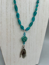 Load image into Gallery viewer, Bring the beauty of nature to your wardrobe with this gorgeous blue-green pressed glass floral beaded necklace. Featuring a large intricate green-blue pressed glass bead, a lucky hand charm, mixed flat matte teal glass beads, and a hook-eye clasp for secure wear, this nature-inspired accessory adds a touch of summer to any outfit. Perfect for day-to-night looks.  Measurements:  Necklace: 19 1/2", Flower and lucky hand pendant 3 1/4"