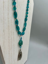 Load image into Gallery viewer, Bring the beauty of nature to your wardrobe with this gorgeous blue-green pressed glass floral beaded necklace. Featuring a large intricate green-blue pressed glass bead, a lucky hand charm, mixed flat matte teal glass beads, and a hook-eye clasp for secure wear, this nature-inspired accessory adds a touch of summer to any outfit. Perfect for day-to-night looks.  Measurements:  Necklace: 19 1/2", Flower and lucky hand pendant 3 1/4"