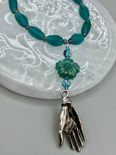 Load image into Gallery viewer, Bring the beauty of nature to your wardrobe with this gorgeous blue-green pressed glass floral beaded necklace. Featuring a large intricate green-blue pressed glass bead, a lucky hand charm, mixed flat matte teal glass beads, and a hook-eye clasp for secure wear, this nature-inspired accessory adds a touch of summer to any outfit. Perfect for day-to-night looks.  Measurements:  Necklace: 19 1/2", Flower and lucky hand pendant 3 1/4"