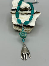 Load image into Gallery viewer, Bring the beauty of nature to your wardrobe with this gorgeous blue-green pressed glass floral beaded necklace. Featuring a large intricate green-blue pressed glass bead, a lucky hand charm, mixed flat matte teal glass beads, and a hook-eye clasp for secure wear, this nature-inspired accessory adds a touch of summer to any outfit. Perfect for day-to-night looks.  Measurements:  Necklace: 19 1/2", Flower and lucky hand pendant 3 1/4"