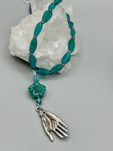 Load image into Gallery viewer, Bring the beauty of nature to your wardrobe with this gorgeous blue-green pressed glass floral beaded necklace. Featuring a large intricate green-blue pressed glass bead, a lucky hand charm, mixed flat matte teal glass beads, and a hook-eye clasp for secure wear, this nature-inspired accessory adds a touch of summer to any outfit. Perfect for day-to-night looks.  Measurements:  Necklace: 19 1/2", Flower and lucky hand pendant 3 1/4"