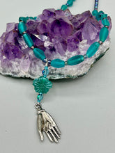 Load image into Gallery viewer, Bring the beauty of nature to your wardrobe with this gorgeous blue-green pressed glass floral beaded necklace. Featuring a large intricate green-blue pressed glass bead, a lucky hand charm, mixed flat matte teal glass beads, and a hook-eye clasp for secure wear, this nature-inspired accessory adds a touch of summer to any outfit. Perfect for day-to-night looks.  Measurements:  Necklace: 19 1/2", Flower and lucky hand pendant 3 1/4"