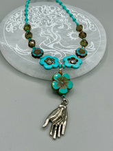Load image into Gallery viewer, Adorn your look and show your affinity for nature with this exquisite handcrafted necklace, composed of a large intricate sea-green hibiscus pressed glass bead, a lucky hand charm, multiple turquoise, and dark avocado green faceted pressed glass beads, and a hook-eye clasp for secure wear. This nature-inspired statement piece brings warmth to any outfit for both day and night. Measurements:  Necklace: 19 1/2", Flower and lucky hand pendant 3"