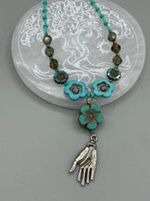 Load image into Gallery viewer, Adorn your look and show your affinity for nature with this exquisite handcrafted necklace, composed of a large intricate sea-green hibiscus pressed glass bead, a lucky hand charm, multiple turquoise, and dark avocado green faceted pressed glass beads, and a hook-eye clasp for secure wear. This nature-inspired statement piece brings warmth to any outfit for both day and night. Measurements:  Necklace: 19 1/2", Flower and lucky hand pendant 3"