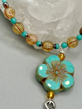Load image into Gallery viewer, Complete your look with this carefully crafted necklace, featuring a large sea-green hibiscus pressed glass bead, a lucky hand charm, assorted champagne melon beads, and turquoise glass beads. All these elements are combined with a hook-eye clasp, ensuring lasting wear. This nature-inspired accessory adds a touch of charm to all kinds of outfits, making it ideal for both day and night. Measurements: Necklace: 18"