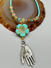 Load image into Gallery viewer, Complete your look with this carefully crafted necklace, featuring a large sea-green hibiscus pressed glass bead, a lucky hand charm, assorted champagne melon beads, and turquoise glass beads. All these elements are combined with a hook-eye clasp, ensuring lasting wear. This nature-inspired accessory adds a touch of charm to all kinds of outfits, making it ideal for both day and night. Measurements: Necklace: 18"