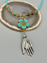Load image into Gallery viewer, Complete your look with this carefully crafted necklace, featuring a large sea-green hibiscus pressed glass bead, a lucky hand charm, assorted champagne melon beads, and turquoise glass beads. All these elements are combined with a hook-eye clasp, ensuring lasting wear. This nature-inspired accessory adds a touch of charm to all kinds of outfits, making it ideal for both day and night. Measurements: Necklace: 18"