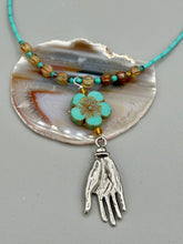Load image into Gallery viewer, Complete your look with this carefully crafted necklace, featuring a large sea-green hibiscus pressed glass bead, a lucky hand charm, assorted champagne melon beads, and turquoise glass beads. All these elements are combined with a hook-eye clasp, ensuring lasting wear. This nature-inspired accessory adds a touch of charm to all kinds of outfits, making it ideal for both day and night. Measurements: Necklace: 18"