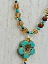 Load image into Gallery viewer, Complete your look with this carefully crafted necklace, featuring a large sea-green hibiscus pressed glass bead, a lucky hand charm, assorted champagne melon beads, and turquoise glass beads. All these elements are combined with a hook-eye clasp, ensuring lasting wear. This nature-inspired accessory adds a touch of charm to all kinds of outfits, making it ideal for both day and night. Measurements: Necklace: 18"