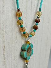 Load image into Gallery viewer, Complete your look with this carefully crafted necklace, featuring a large sea-green hibiscus pressed glass bead, a lucky hand charm, assorted champagne melon beads, and turquoise glass beads. All these elements are combined with a hook-eye clasp, ensuring lasting wear. This nature-inspired accessory adds a touch of charm to all kinds of outfits, making it ideal for both day and night. Measurements: Necklace: 18"