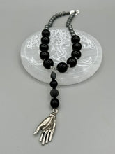 Load image into Gallery viewer, This elegant hematite and black glass beaded necklace is the perfect accessory for your unique look. With a large black and gray glass beads adorned with a lucky hand charm, it exudes a dark, alluring vibe. Complemented with floral shaped metal beads, it will make a bold statement.  Measurements:  Necklace: 18” in length - lucky hand charm is 3 1 /2” Height