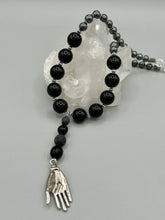 Load image into Gallery viewer, This elegant hematite and black glass beaded necklace is the perfect accessory for your unique look. With a large black and gray glass beads adorned with a lucky hand charm, it exudes a dark, alluring vibe. Complemented with floral shaped metal beads, it will make a bold statement.  Measurements:  Necklace: 18” in length - lucky hand charm is 3 1 /2” Height