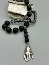 Load image into Gallery viewer, This elegant hematite and black glass beaded necklace is the perfect accessory for your unique look. With a large black and gray glass beads adorned with a lucky hand charm, it exudes a dark, alluring vibe. Complemented with floral shaped metal beads, it will make a bold statement.  Measurements:  Necklace: 18” in length - lucky hand charm is 3 1 /2” Height