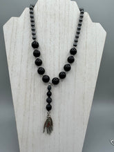 Load image into Gallery viewer, This elegant hematite and black glass beaded necklace is the perfect accessory for your unique look. With a large black and gray glass beads adorned with a lucky hand charm, it exudes a dark, alluring vibe. Complemented with floral shaped metal beads, it will make a bold statement.  Measurements:  Necklace: 18” in length - lucky hand charm is 3 1 /2” Height