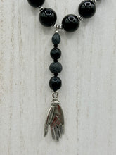 Load image into Gallery viewer, This elegant hematite and black glass beaded necklace is the perfect accessory for your unique look. With a large black and gray glass beads adorned with a lucky hand charm, it exudes a dark, alluring vibe. Complemented with floral shaped metal beads, it will make a bold statement.  Measurements:  Necklace: 18” in length - lucky hand charm is 3 1 /2” Height