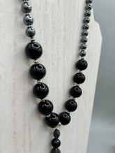 Load image into Gallery viewer, This elegant hematite and black glass beaded necklace is the perfect accessory for your unique look. With a large black and gray glass beads adorned with a lucky hand charm, it exudes a dark, alluring vibe. Complemented with floral shaped metal beads, it will make a bold statement.  Measurements:  Necklace: 18” in length - lucky hand charm is 3 1 /2” Height