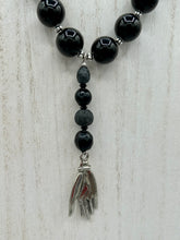 Load image into Gallery viewer, This elegant hematite and black glass beaded necklace is the perfect accessory for your unique look. With a large black and gray glass beads adorned with a lucky hand charm, it exudes a dark, alluring vibe. Complemented with floral shaped metal beads, it will make a bold statement.  Measurements:  Necklace: 18” in length - lucky hand charm is 3 1 /2” Height