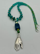 Load image into Gallery viewer, Bring the beauty of nature to your wardrobe with this gorgeous blue-green glass beaded necklace. Featuring a large rectangular blue-green glass bead, a lucky hand charm, mixed flat matte teal glass beads, and a hook-eye clasp for secure wear, this nature-inspired accessory adds a touch of summer to any outfit. Perfect for day-to-night looks.   Measurements:  Necklace: 19", Lucky hand pendant 3 1/2"